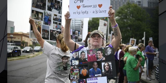 In this Aug. 17, 2018, file photo, Lynn Wencus of Wrentham, Mass., holds a sign with a picture of her son Jeff and wears a sign of others' loved ones lost to OxyContin and other opioids during a protest at Purdue Pharma LLP headquarters in Stamford, Conn. (AP Photo/Jessica Hill, File)