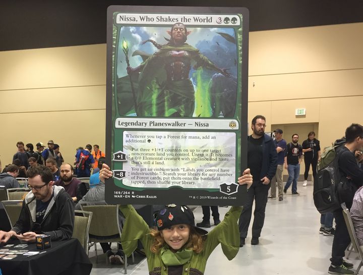 Dana holding up an oversized card showing Nissa, a legendary figure within the game.