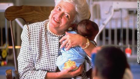 First Lady Barbara Bush holding baby while two-year-old child takes a photo wtih a toy camera at hospice for children with  AIDS. 
